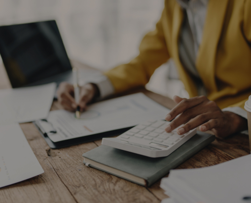 A person in a yellow blazer calculates HVAC service costs on a clipboard at a desk with papers, laptop, and coffee in British Columbia.