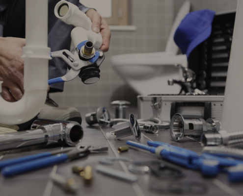 Plumber repairing white bathroom pipes with a wrench, tools and parts nearby—ideal for BC HVAC service pages. Toilet in background.