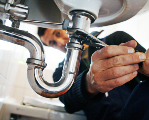 Expert plumber in BC using a wrench to repair bathroom sink pipes, focusing on hands and plumbing details for HVAC services.