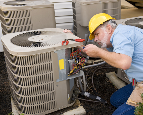 HVAC technician in yellow hard hat repairs outdoor AC unit, inspecting wires. Multiple air conditioning units on site in British Columbia.