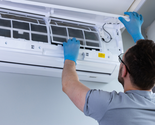 An HVAC technician in blue gloves cleans an air conditioner, lifting the panel to access filters in a British Columbia home.