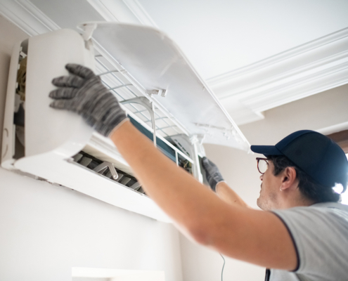 A technician in gloves and a cap checks a wall-mounted ductless mini-split AC, lifting the panel for HVAC service in British Columbia.