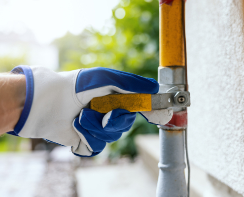 A gloved hand adjusts a yellow gas line valve outside a home in BC, near a white wall and green plants—HVAC service in action.