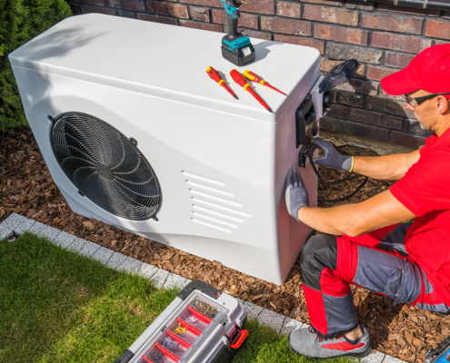 HVAC technician in red uniform services outdoor heat pump beside a brick wall and grass, showcasing expert BC heat pump repair.