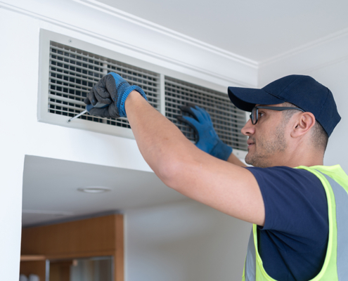 An HVAC technician in safety gear services an indoor air vent with a screwdriver to improve air quality in a British Columbia home.