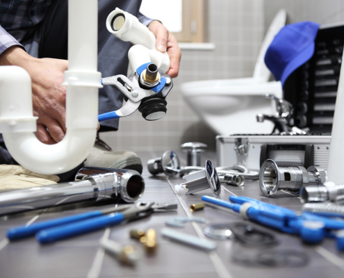 A plumber in a bathroom uses a wrench on a white pipe under the sink, surrounded by plumbing tools—HVAC service in BC.