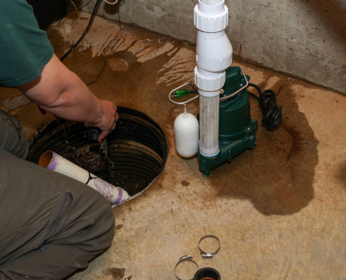 HVAC technician in a British Columbia basement repairs a sump pump, hand in pit, with pipes, water, and tools on the concrete floor.