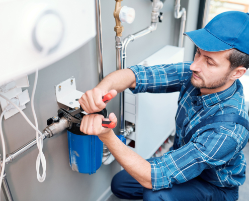 Plumber in blue cap and plaid shirt using a wrench to service a water filter, pipes and outlets visible—HVAC in British Columbia.