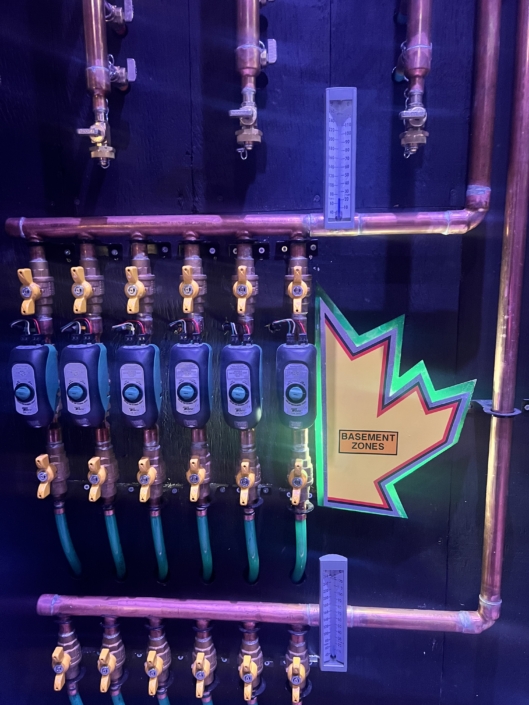 Copper pipes, valves, and gauges on a wall under purple-blue light, with a BASEMENT ZONES leaf sign—HVAC setup in BC.