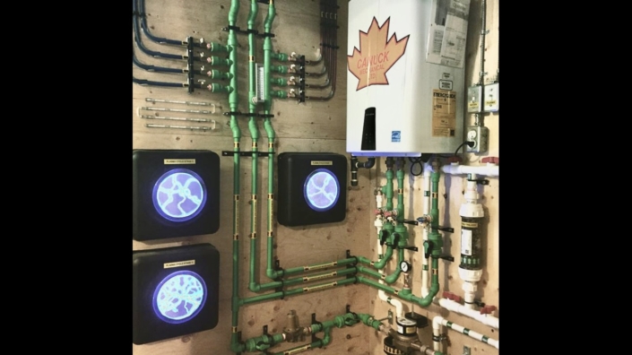 Organized HVAC utility wall in BC featuring green pipes, a tankless water heater, blue fan units, valves, and meters.