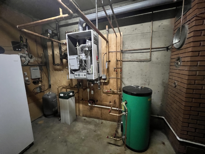 Utility room in BC with exposed pipes, boiler or furnace, green water tank, white HVAC appliance, concrete floor and brick wall.