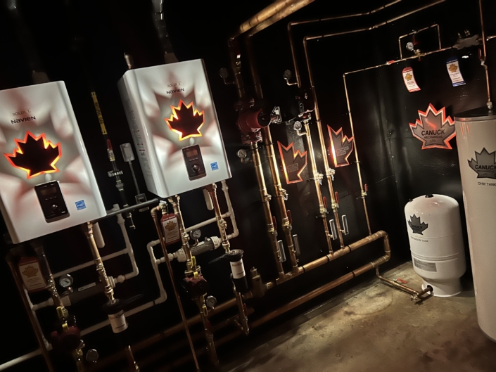 Dimly lit room with several water heaters and tanks, CANUCK maple leaf logos highlight Canadian HVAC services in British Columbia.
