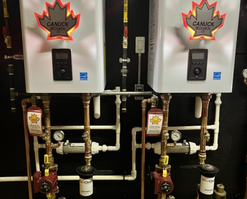 Two CANUCK Navien boilers with maple leaf logos are installed side by side on a black wall, featuring copper pipes and HVAC gauges.