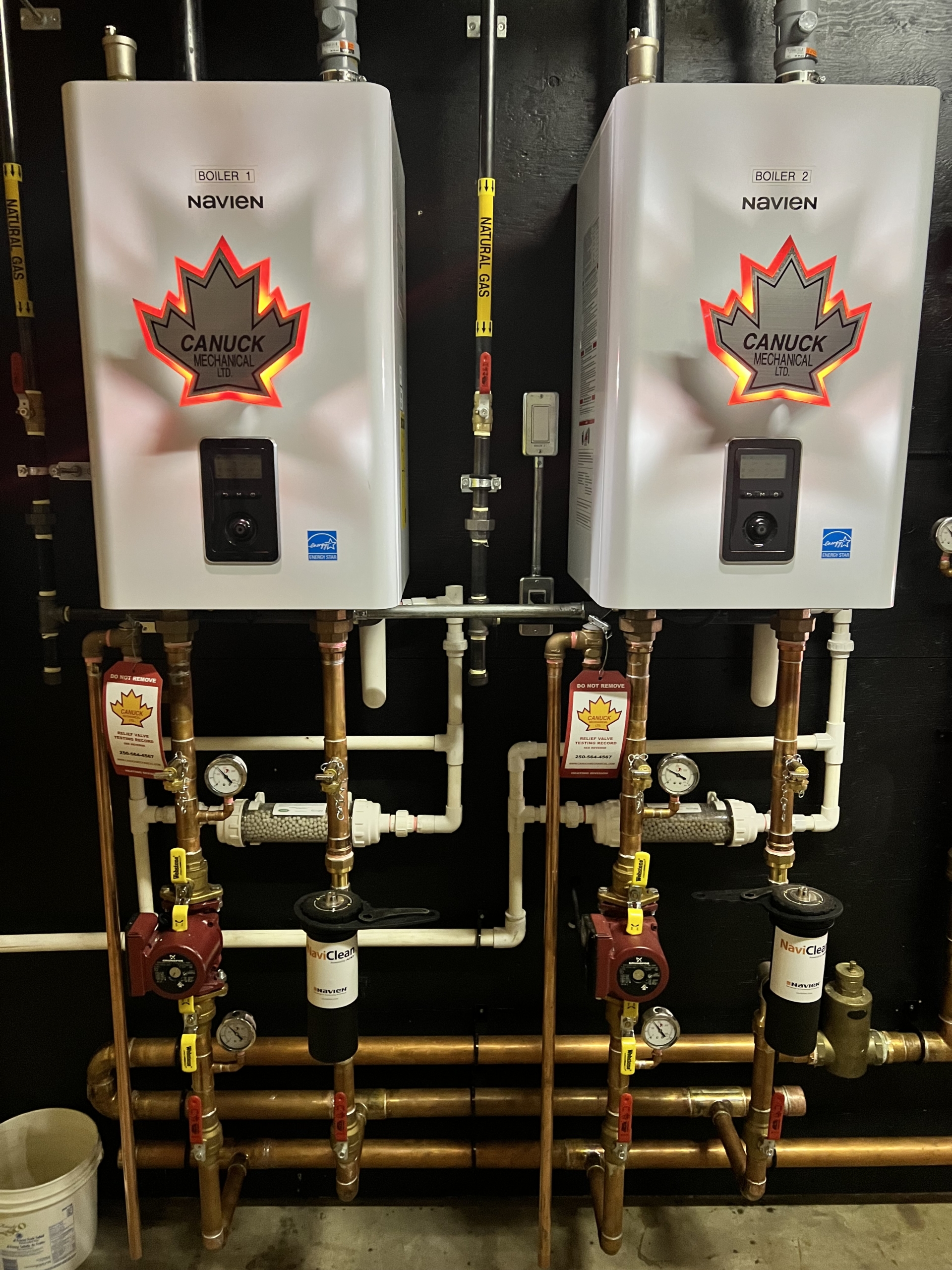 Two CANUCK Navien boilers with maple leaf logos are installed side by side on a black wall, featuring copper pipes and HVAC gauges.
