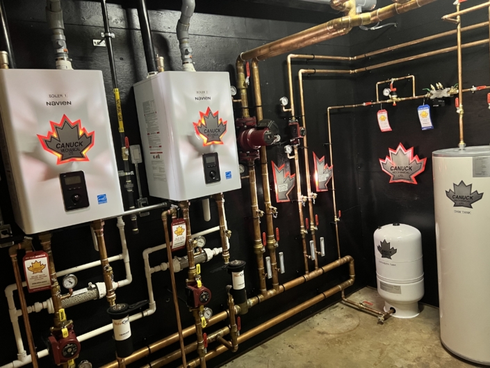 Mechanical room with two white Navien boilers, copper pipes, gauges, and a tank—Canuck Plumbing HVAC services in BC.