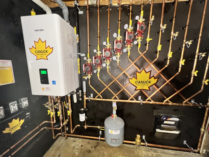 A modern wall-mounted boiler system with copper pipes, red valves, gauges, and a gray tank, labeled CANUCK MECHANICAL LTD.—HVAC BC.