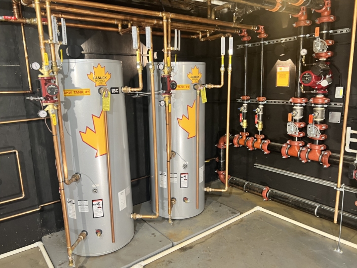 Two silver water heaters with maple leaf decals, copper pipes, and valves in a spotless BC mechanical room—HVAC services shown.